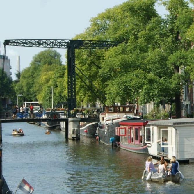 Authentic Amsterdam canal scene with boats cruising past houseboats and an industrial bridge in the background.
